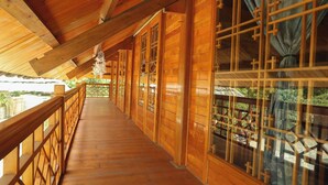 Un balcón de madera con barandilla, amplias ventanas con cortinas y vistas al verdor.