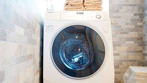 A front-loading washing machine with a control panel, towels stacked on top, and a tiled wall. - Mcity in Furujima (Naha)