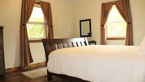 A bedroom with a wooden bed, beige walls, and two windows with brown curtains. - Trailhead Suites (Marysville)