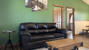 A living room with a black leather sofa, a wooden coffee table with wheels, a small side table, and a green wall with three framed pictures. - Trailhead Suites (Marysville)