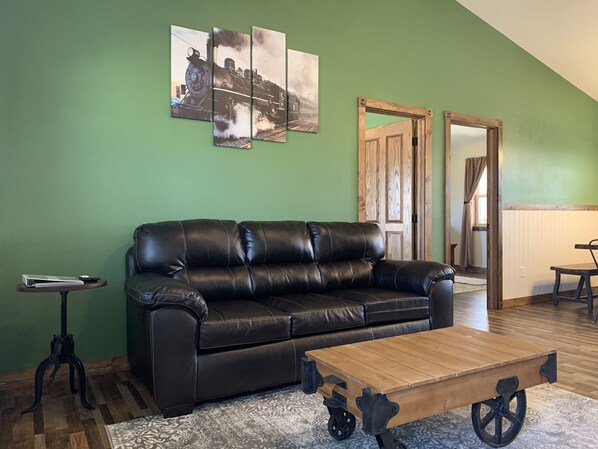 Una sala de estar con un sofá de cuero negro, una mesa de centro de madera con ruedas, una mesita auxiliar y una pared verde con tres cuadros enmarcados.