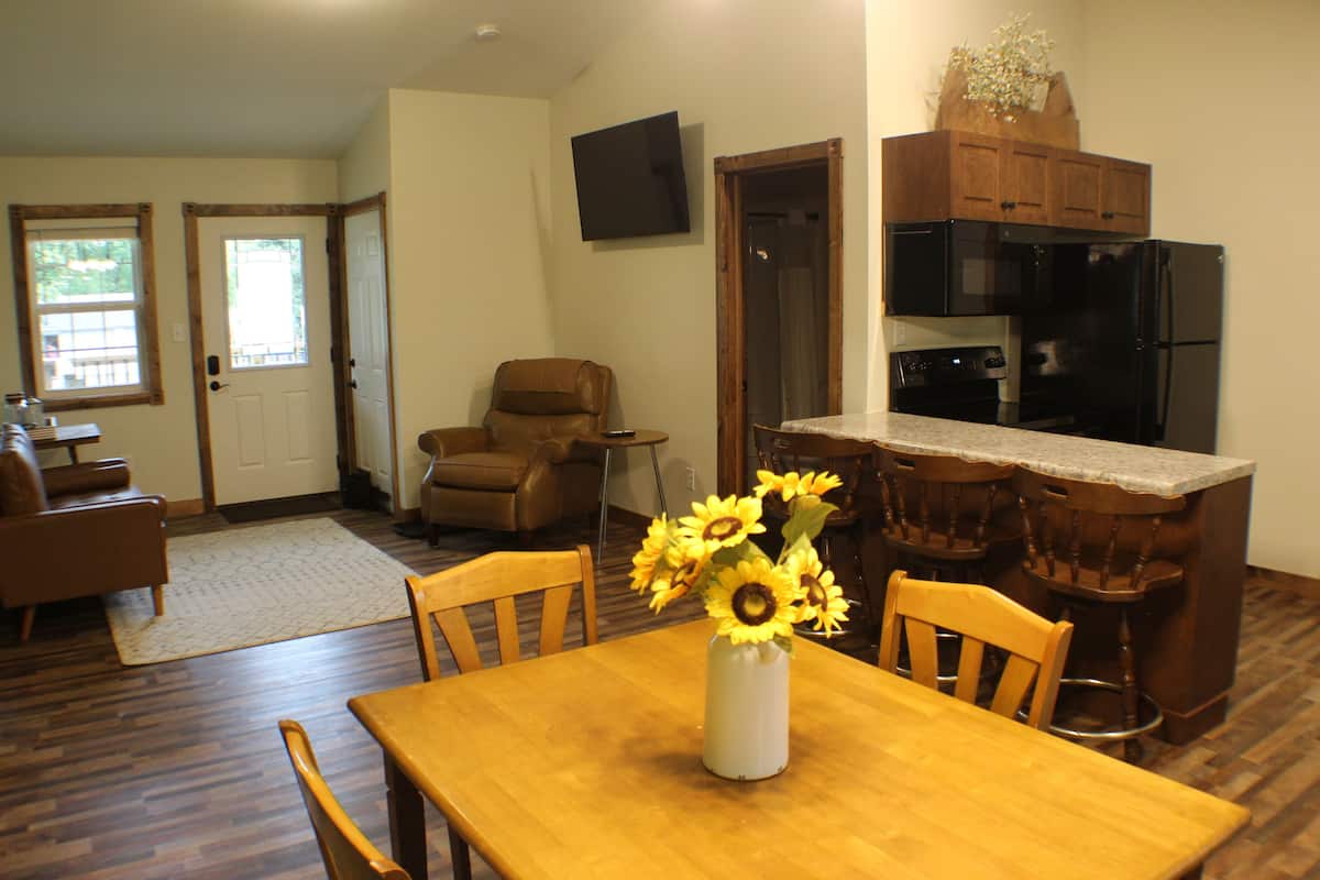 A dining area with a wooden table and chairs, a vase of sunflowers, and a kitchen with a black refrigerator and wooden cabinets.