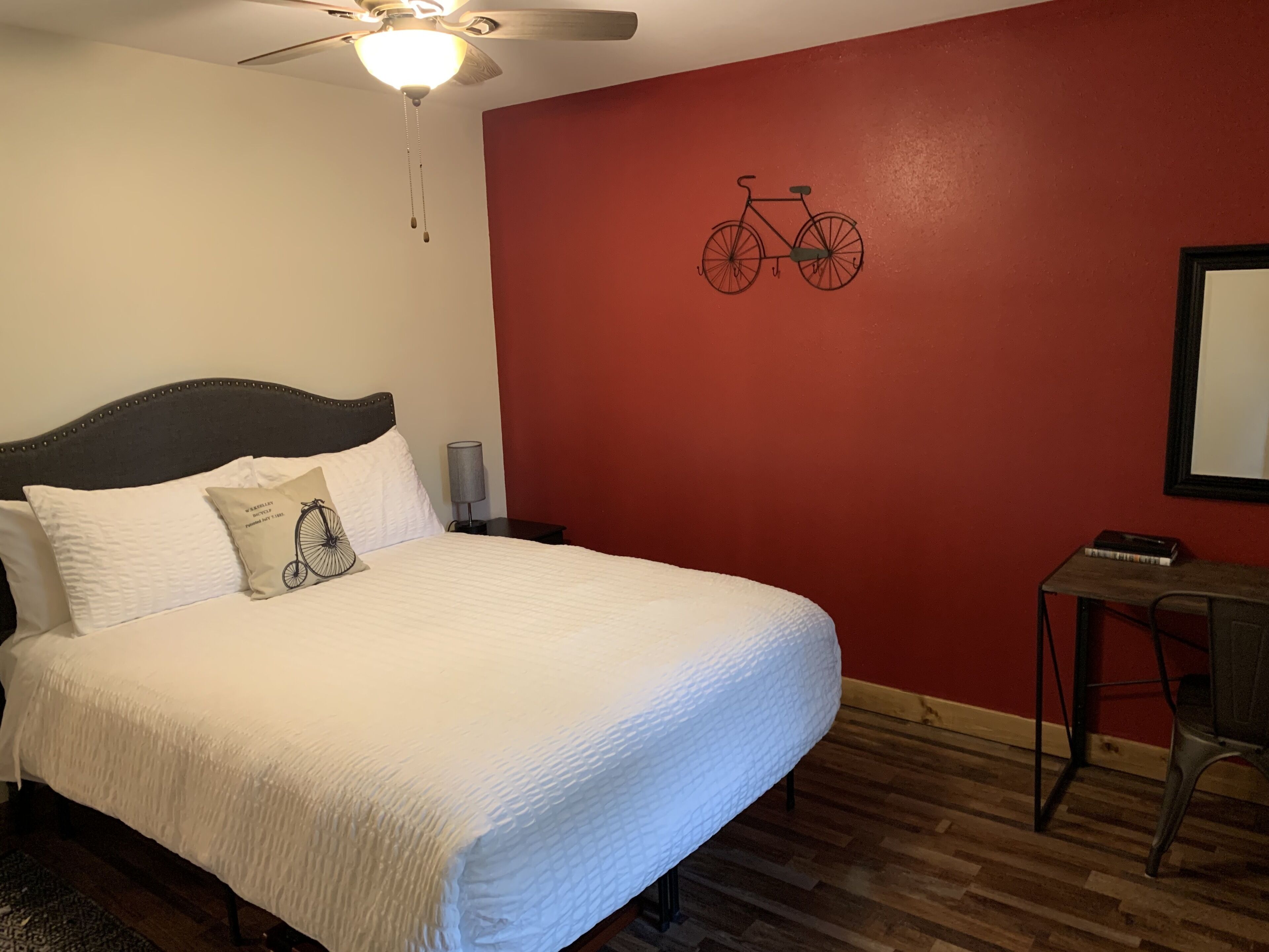 A bedroom with a bed, a ceiling fan, a wall-mounted bicycle illustration, a desk, and a chair.