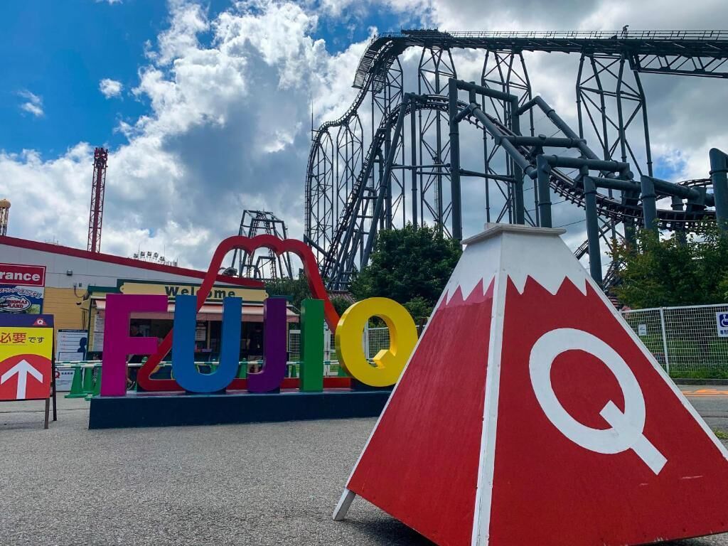 Una montaña rusa, una gran señal roja y blanca con la letra Q, y letras de colores que deletrean "FUTAGO" en la entrada de un parque temático.