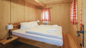 Een houten kamer met een bed, een nachtkastje met een lamp en een raam met een rood-wit geruit gordijn.