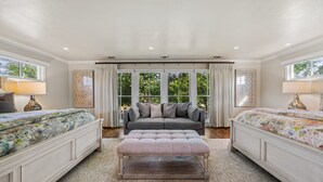 A spacious bedroom with a large bed, a sofa, and a bench. There are two windows with curtains, and a view of greenery outside. - The Live Oak Haus (Sleeps 19) - 6 Bed | 6 Bath | Pool + Hot Tub + Fire Pit (Fredericksburg)