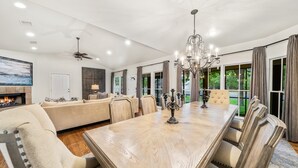 A spacious living room with a large wooden dining table, comfortable seating, a fireplace, and large windows offering a view of the outdoors. - The Live Oak Haus (Sleeps 19) - 6 Bed | 6 Bath | Pool + Hot Tub + Fire Pit (Fredericksburg)