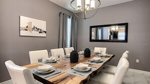 Een eetkamer met een lange houten tafel gedekt voor zes personen, witte stoelen, een kroonluchter, een stadsgezicht schilderij en een spiegel aan de muur.
