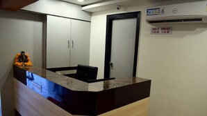 A reception desk with a sign indicating check-in and check-out times, a black chair, and a statue of a deity on the left. - Hotel Palak Palace (Ahmedabad)