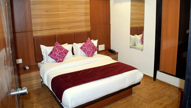 A hotel room with a wooden headboard, a large bed with white bedding and red accents, a bedside table with a phone, and a ceiling fan. - Hotel Palak Palace (Ahmedabad)