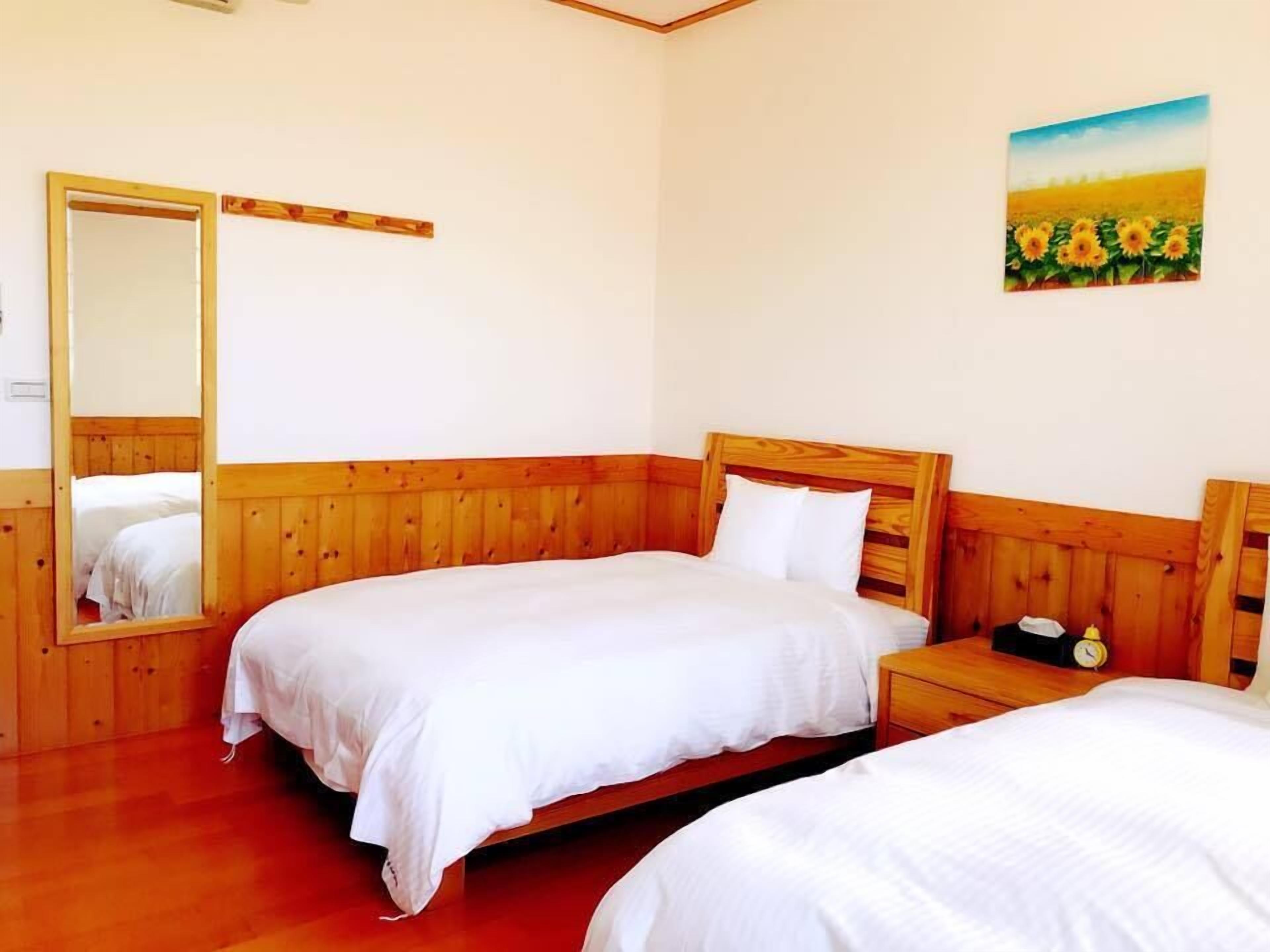A room with two wooden beds, a wooden nightstand with a clock, and a painting of sunflowers on the wall.