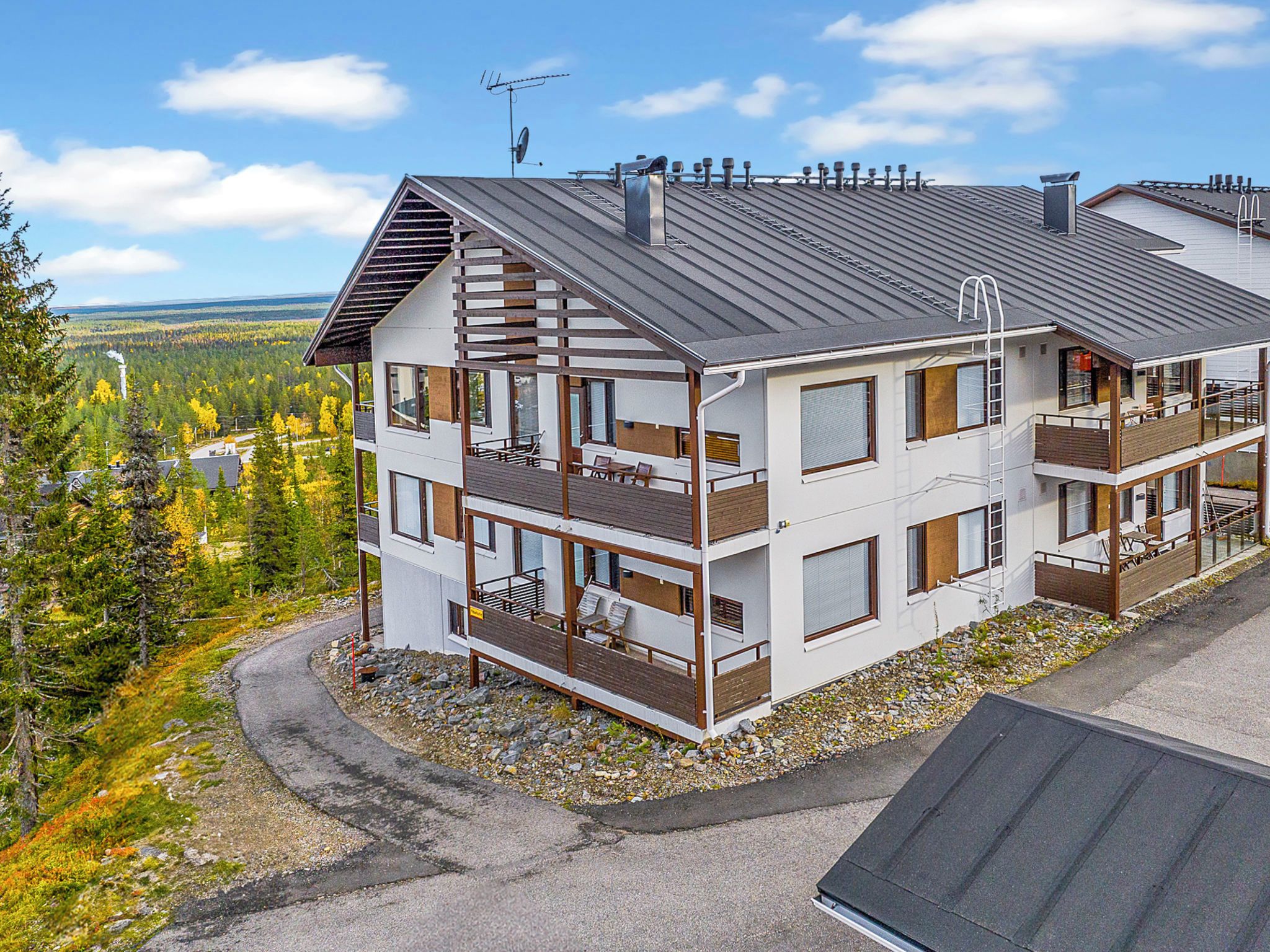 A multi-story building with balconies, a sloped roof, and a view of a forested area.