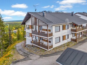 A multi-story building with balconies, a sloped roof, and a view of a forested area.