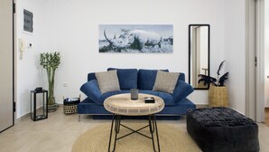 A modern living room with a blue sofa, a round wicker table, a black ottoman, and a large mirror. - Modern Bohemian Style Sun-Splashed Loft (Athens)