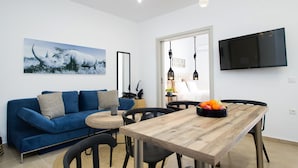 A modern living room with a blue sofa, a wooden dining table, and a flat-screen TV mounted on the wall. - Modern Bohemian Style Sun-Splashed Loft (Athens)