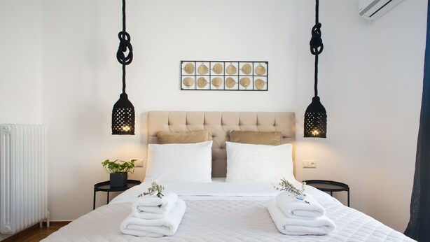 A neatly made bed with white linens and two towels, a tufted headboard, and a wall-mounted decorative piece above. - Modern Bohemian Style Sun-Splashed Loft (Athens)