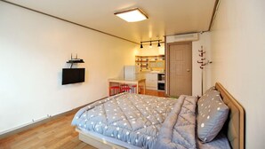 A bedroom with a bed, a desk with chairs, a TV, and a kitchenette.