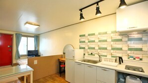 A compact kitchen with a sink, a refrigerator, and a microwave. There is a window with curtains, a small table, and a chair.
