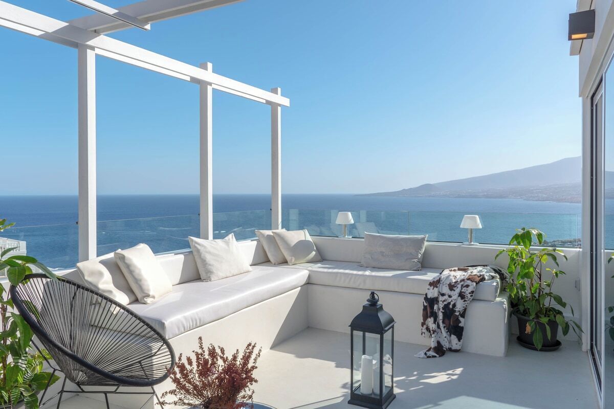 A balcony with a white sofa, a chair, a potted plant, a lantern, and a view of the sea.
