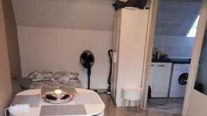 Pieni makuuhuone, jossa on sänky, yöpöytä ja pieni työpöytä tuolilla. Seinälle on asennettu tuuletin, ja viereisessä huoneessa näkyy pesukone.