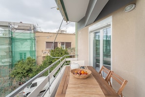 A balcony with a wooden table, a wicker basket with fruits, and a view of a building with a green fence and trees. - Sun splashed sanctum balconies Acropolis (Athens)