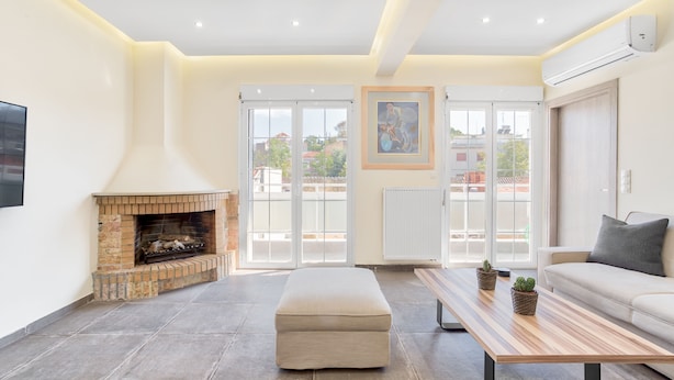 A modern living room with a fireplace, a sofa, a coffee table, and a large window with a view of buildings. - Serene Acropolis View Roofdeck (Athens)