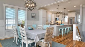 A modern kitchen with white cabinetry, a dining area with a table and chairs, and a bar area with stools. - Anita Getaway: Charming 4-bedroom home in Oak Island with ocean views and private pool. (Oak Island)