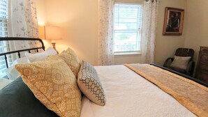 A neatly made bed with patterned pillows, a bedside lamp, and a window with blinds. - Chickadees Cottage and Pottery - centrally located, private comfortable cottage. (Arden)
