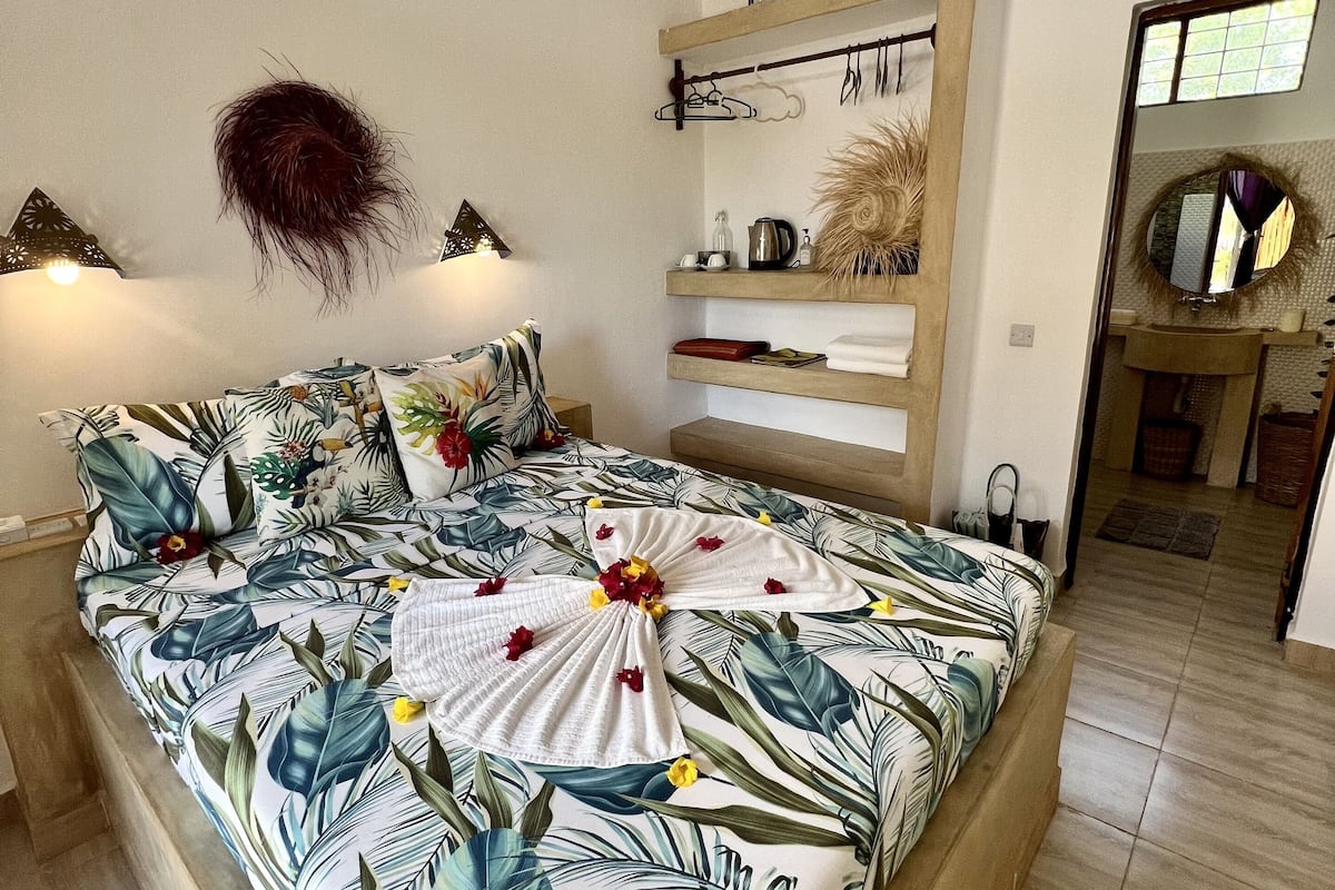 Un dormitori amb un llit, un ventilador de sostre, una estanteria penjada a la paret amb objectes decoratius i una porta que dona a una altra habitació.