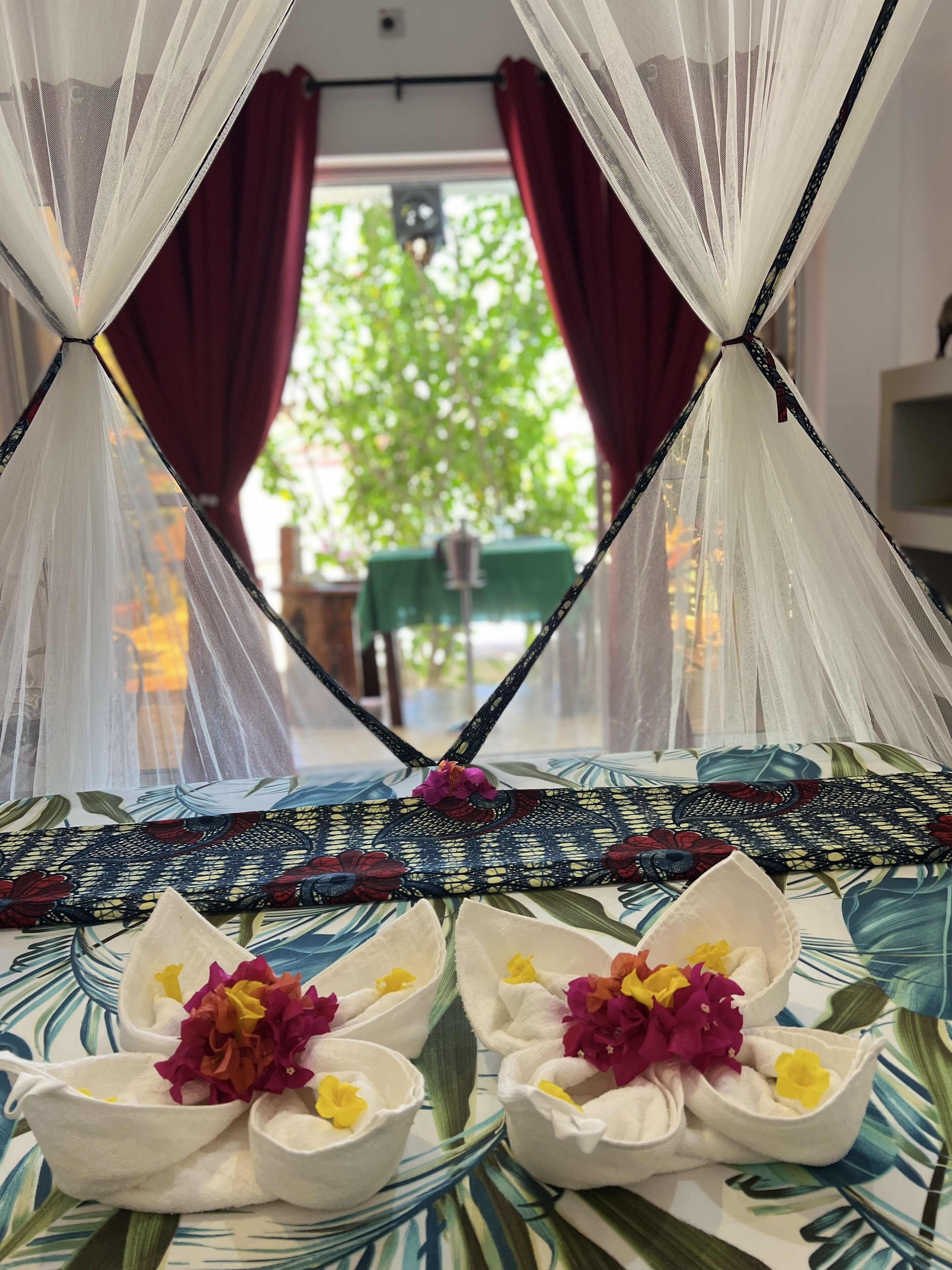 Una habitación con una cama cubierta con una colcha de motivos tropicales, un mosquitero y arreglos florales decorativos sobre los almohadones.