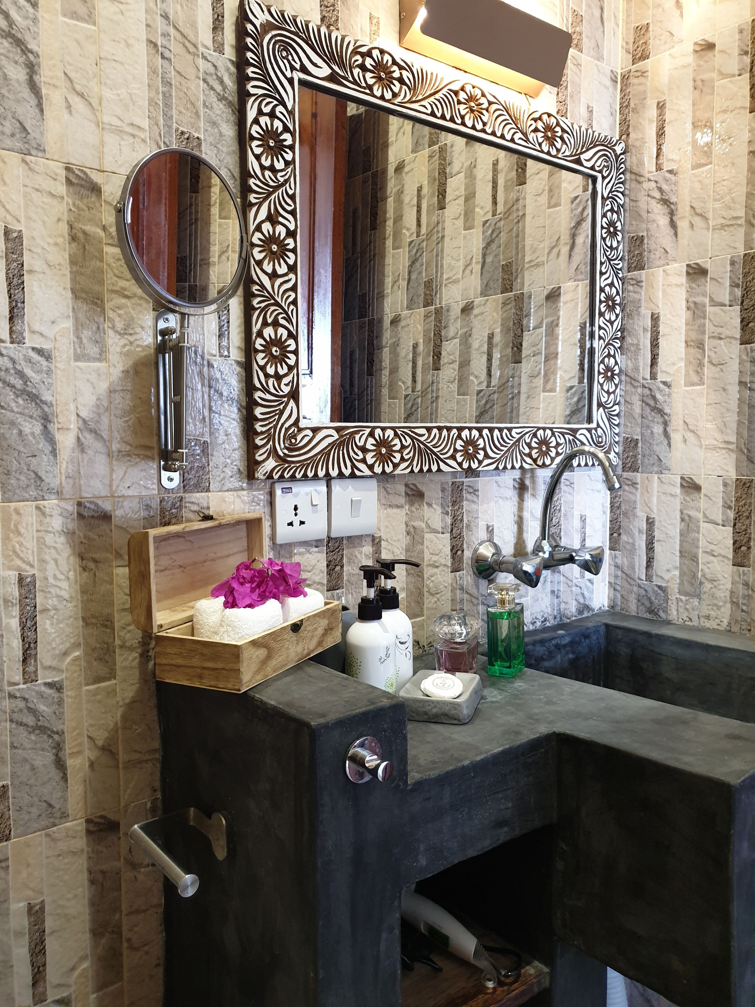 Un baño con pared de mármol, tocador de madera, un espejo grande con marco decorativo, un espejo redondo, una caja de madera con flores y un lavamanos negro.