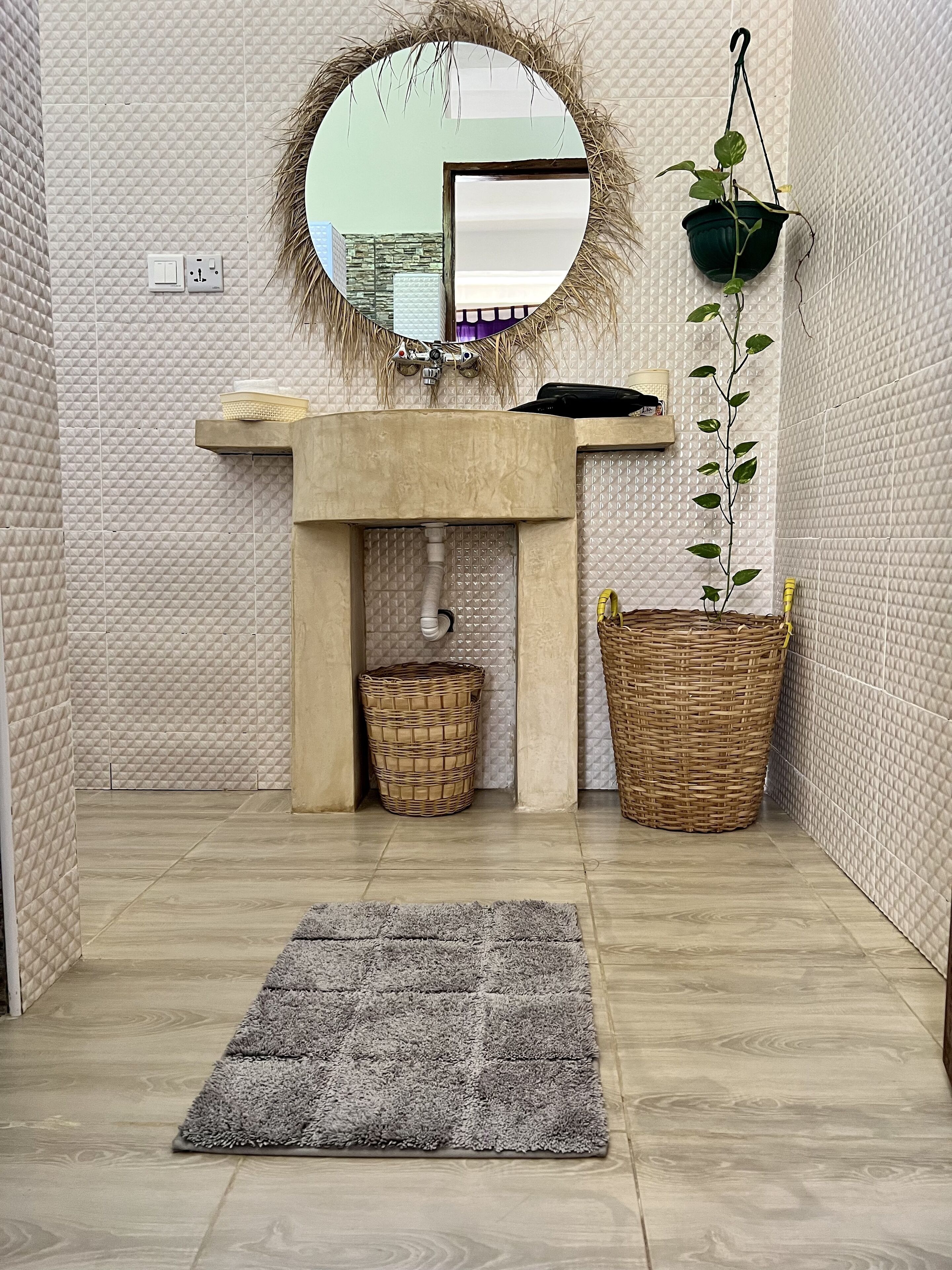 Une salle de bain avec un miroir rond, un lavabo, une plante suspendue, un panier en osier et un tapis gris.