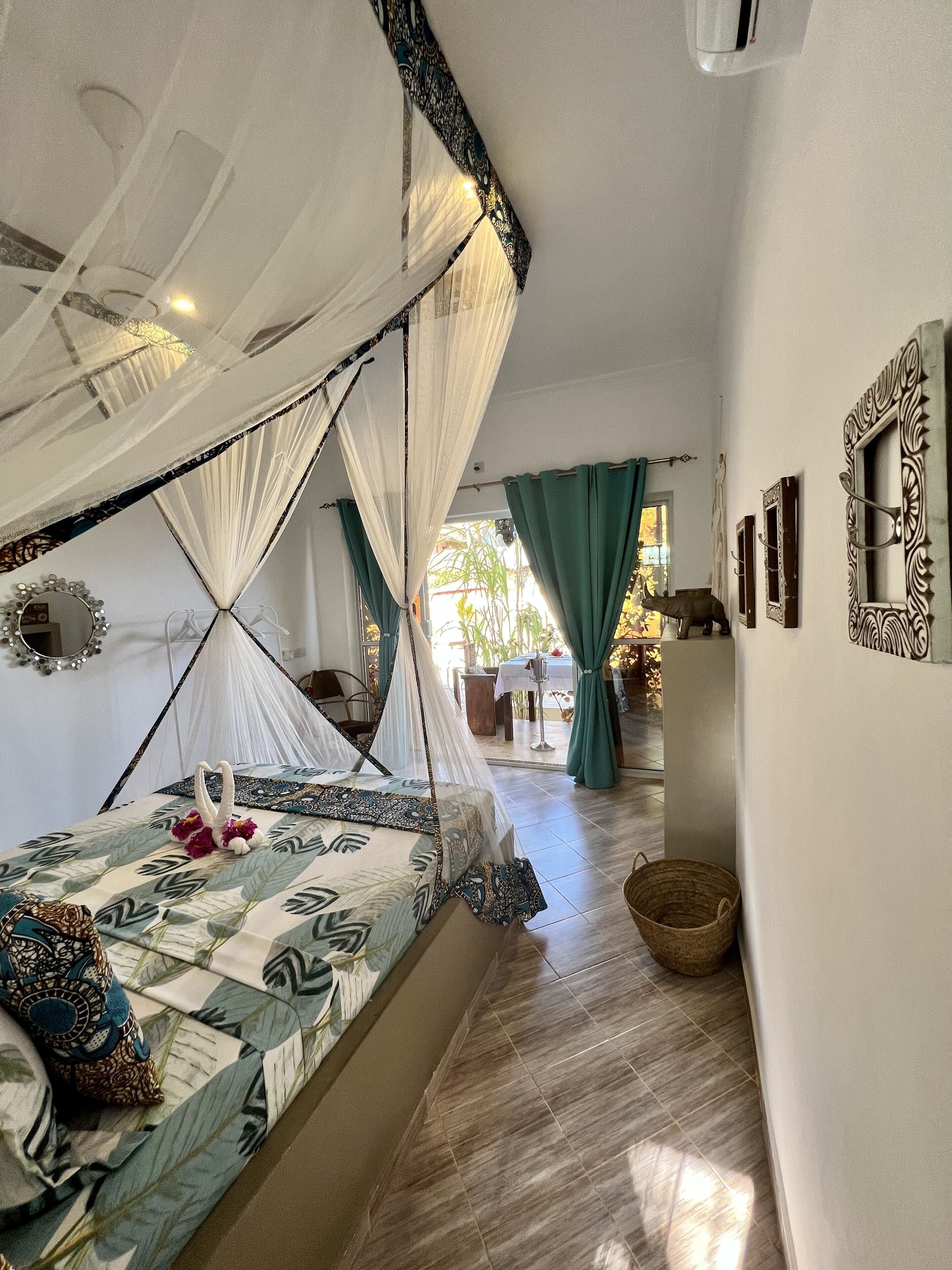 A room with a bed under a mosquito net, a small table with a vase, and a view of the outside.