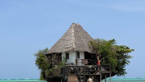 A thatched-roof bungalow on stilts over turquoise water, with a person standing on the deck. - Gadea Boutique Hotel (Jambiani)
