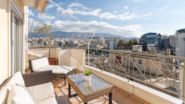 A balcony with wicker furniture, a potted plant, and a clear view of the cityscape and mountains in the background. - Penthouse w Acropolis View & Parking (Athens)