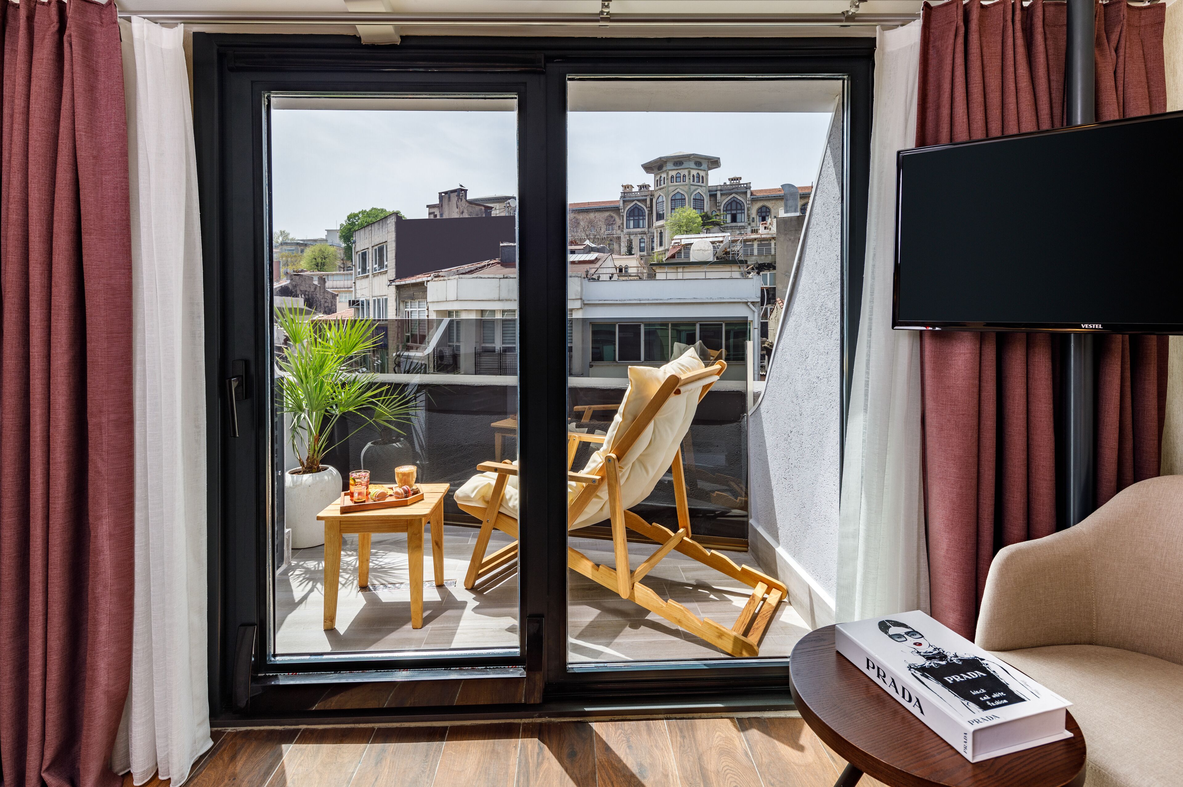 Una habitación con puerta de vidrio corredera que da a un balcón, un televisor de pantalla plana montado en la pared, un sofá color beige, una silla de madera, una mesita con una planta y un libro titulado "PRADA".