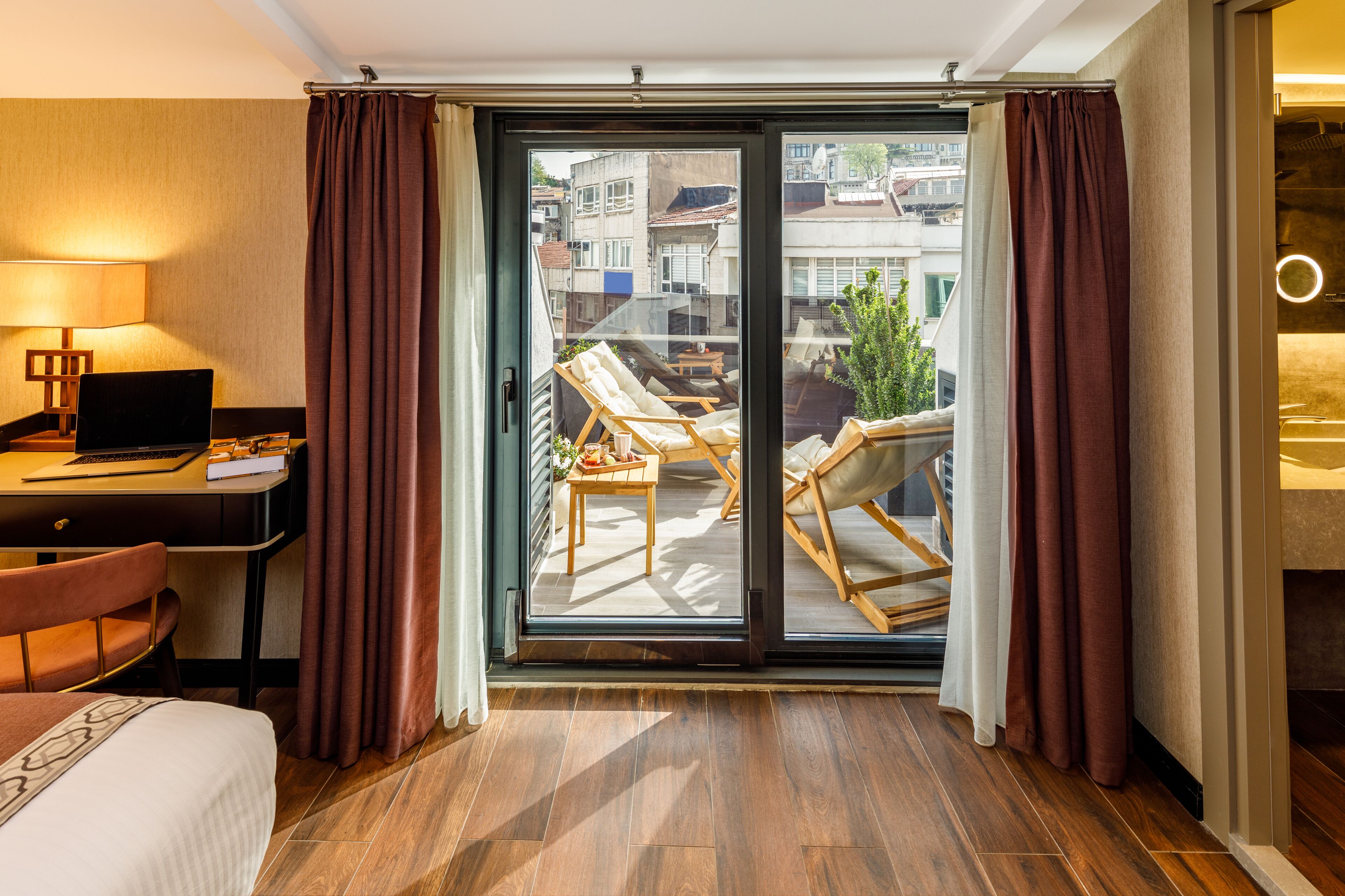 Ein Hotelzimmer mit Bett, Schreibtisch und Computer, ein Balkon mit Stühlen und Tisch sowie Blick auf Gebäude.