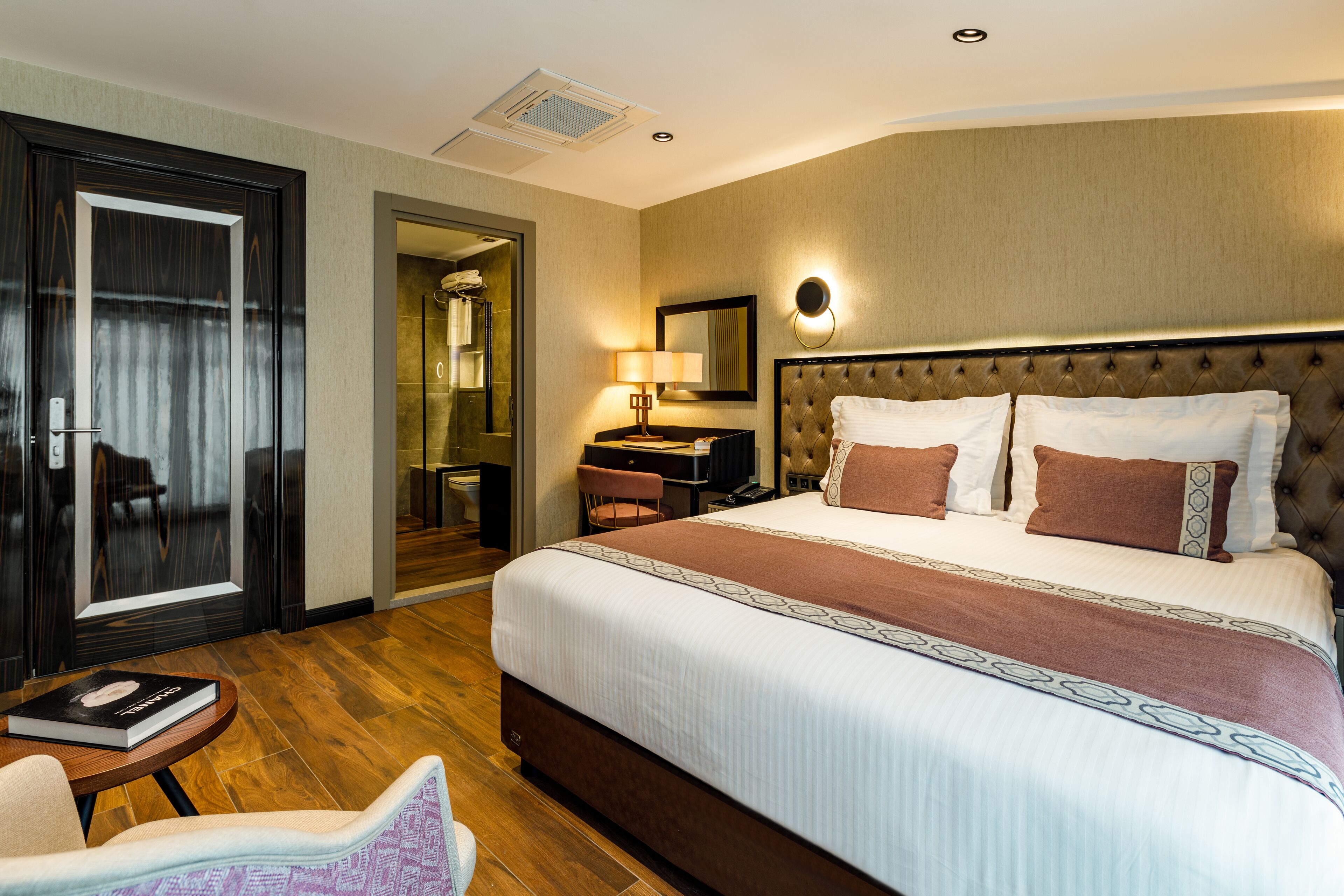 A hotel room with a large bed, a desk with a chair, a small table, and a door leading to a bathroom.