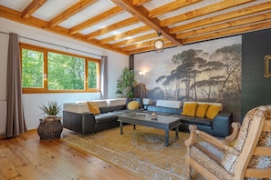 A room with a wooden ceiling, large windows, a sofa set, a patterned rug, and a painting on the wall.