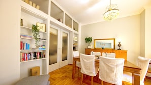 A dining area with a wooden table and chairs, a bookshelf with books, a plant, and a chandelier. - Your home under  Acropolis Central& safe (Athens)