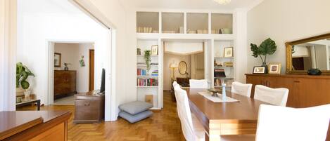 A spacious dining area with a wooden table and white chairs, flanked by white shelving units and a large mirror.