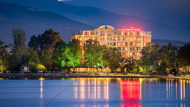A hotel with a prominent sign, surrounded by trees and a calm body of water reflecting the building and sky. - Landmark Creek Hotel & Wellness (Plovdiv)