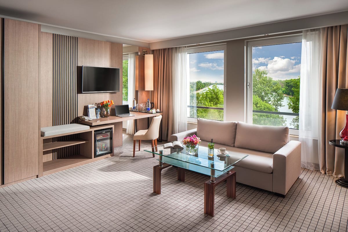 A modern hotel room with a glass coffee table, a sofa, a desk with a computer, a TV, and a balcony with a scenic view.