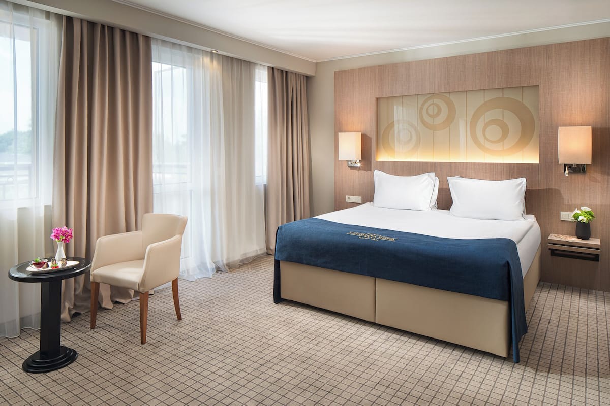 A hotel room with a large bed, a chair, a small table with flowers, and a wall-mounted lamp.