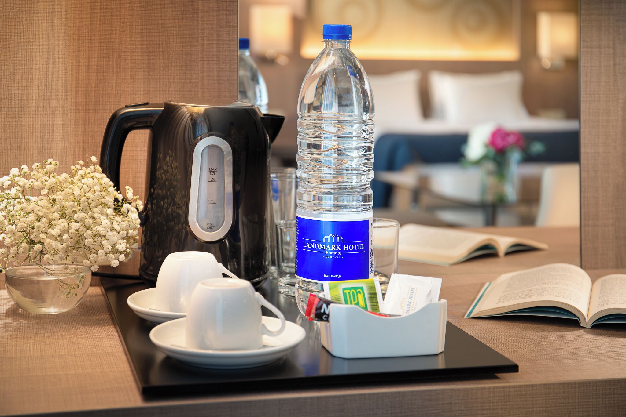 Ein Hotelzimmer mit Kaffeemaschine, einer Wasserflasche, einer Blumenschale, einer Tasse, einem Buch und einem Tablett mit Taschentüchern.