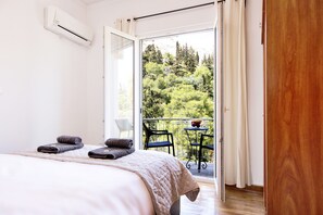A bedroom with a bed, two rolled towels, a balcony with a table and chairs, and a view of greenery. - Acropolis Hillside Escape - Sea & Nature View (Athens)