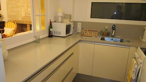 A compact kitchen with a microwave, a sink, and a window overlooking a room with a chair and a plant. - Exceptional location, 1st line of the sea, 86m2 apartment with terrace (Sant Antoni de Calonge)