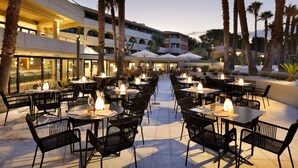 An outdoor dining area with tables and chairs, surrounded by palm trees and a building with multiple balconies. - The Signature Level at Grand Palladium Sicilia Resort & Spa (Campofelice di Roccella)