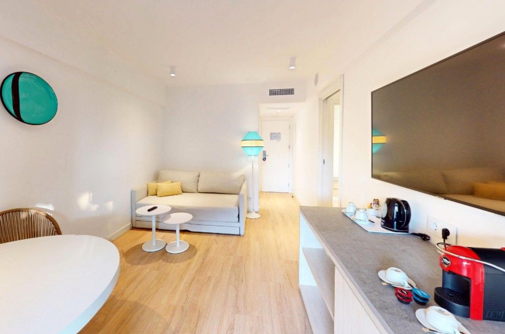 A modern hotel room with a sofa, a small round table, a large mirror, and a kitchenette area with a coffee machine and tea set.