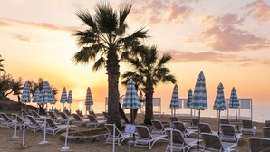 A beach with rows of sun loungers and striped umbrellas, palm trees, and a sunset in the background. - The Signature Level at Grand Palladium Sicilia Resort & Spa (Campofelice di Roccella)
